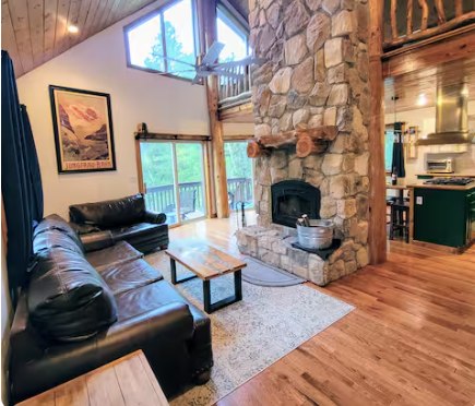 Mountain House great room with stone fireplace, leather sofas, and tall windows looking out to the trees